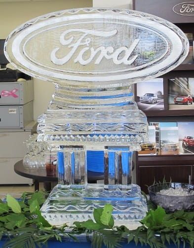 Glass Ford logo sculpture displayed indoors with greenery in the foreground