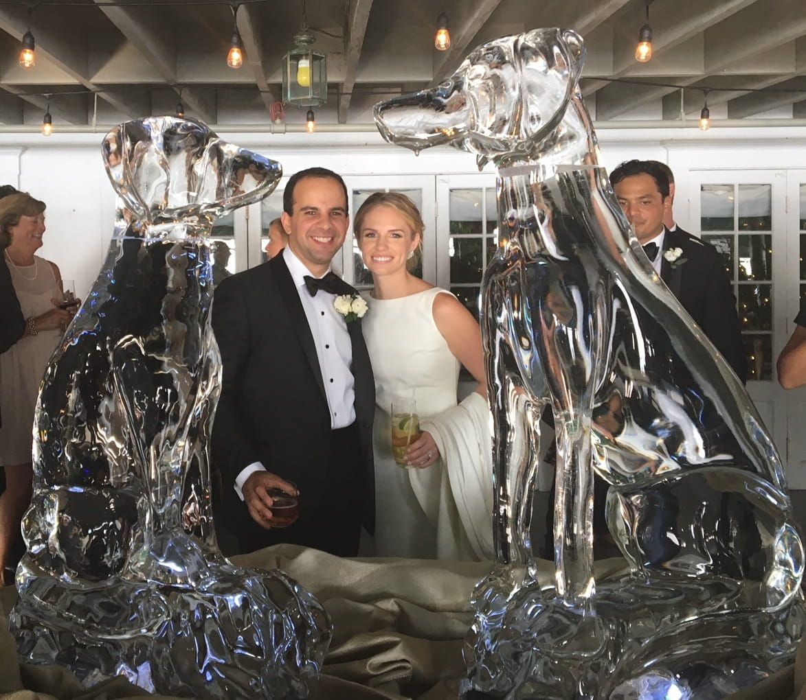 Couple posing among large clear ice sculptures at an indoor event, smiling in formal attire.