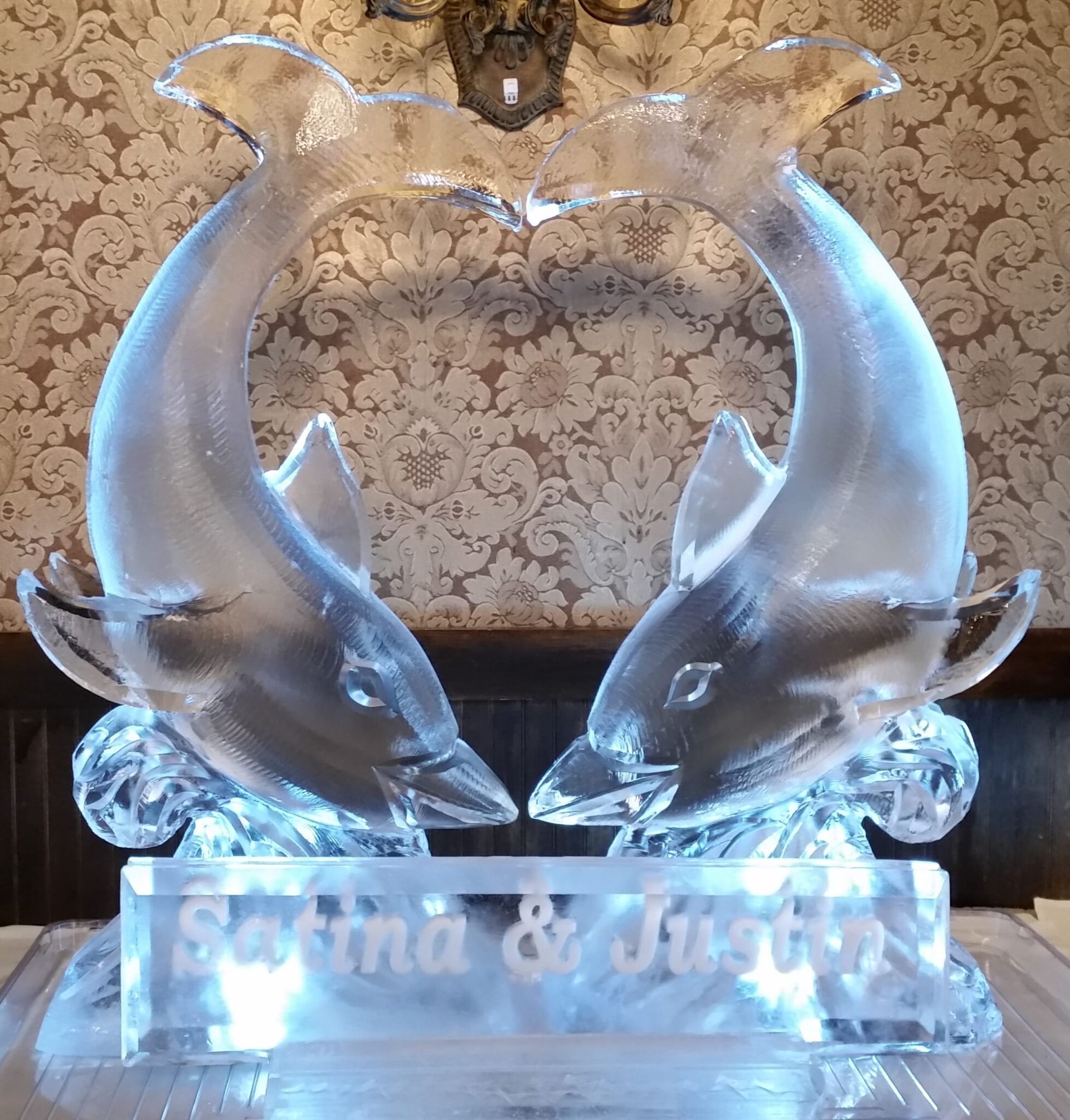 Two glass dolphins arcing around a heart-shaped center on a lit crystal base with “Fatima & Just…” engraved.