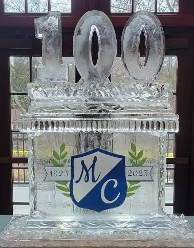 Ice sculpture with “100” and a blue shield logo in front of windows, flanked by “1923” and “2023” marks.
