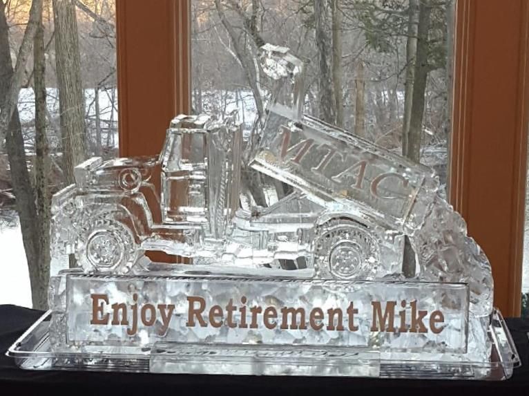 Clear glass construction sign reading “Enjoy Retirement Mike” with a truck and excavator on a windowsill