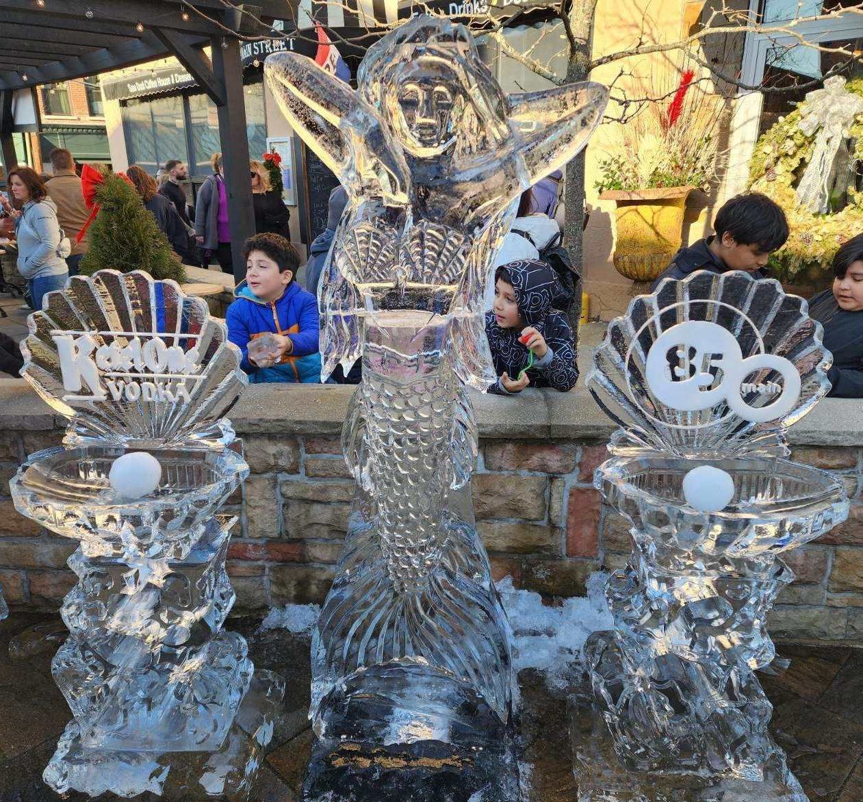 Ice sculpture display with a central mermaid and two side pedestals in a festive outdoor setting, with people behind.