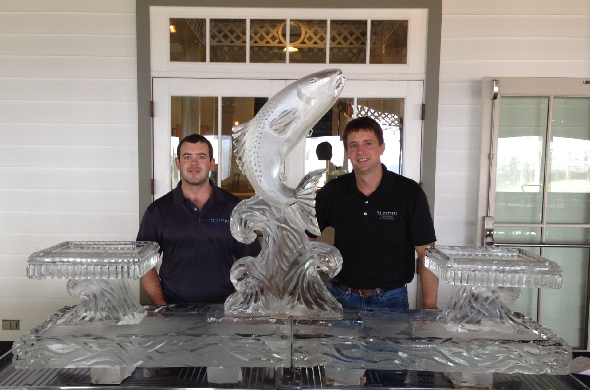Two men standing behind an ornate silver ice sculpture on a table in front of a building entrance
