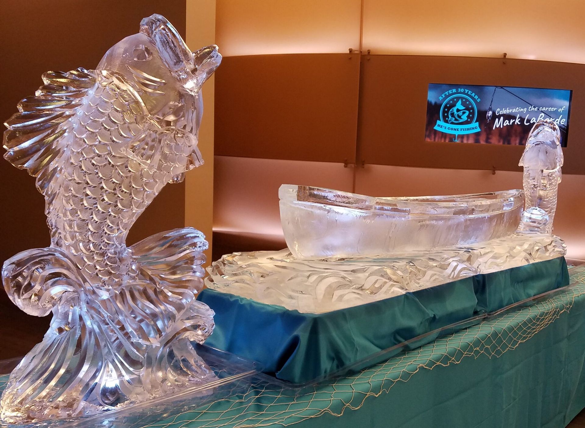 Ice sculpture of a fish beside a small ice boat on a teal-covered table in a banquet hall