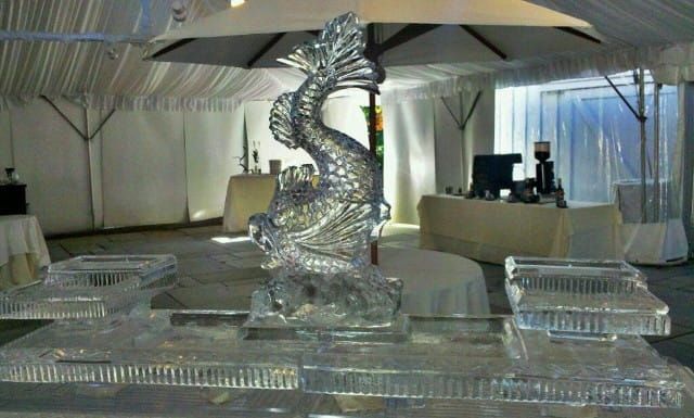 Silver ice sculpture of a dragon beneath umbrellas on a reflective platform at an indoor event