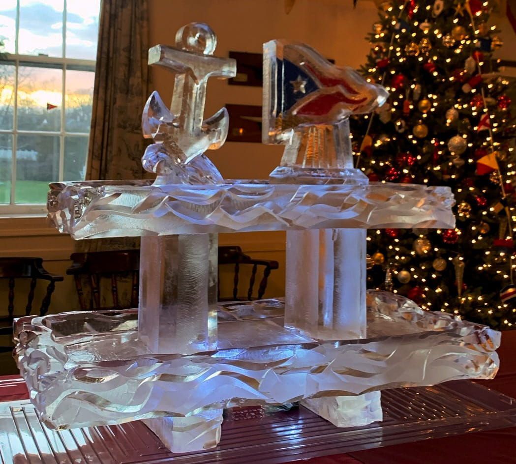 Ice sculpture with an anchor and flags on a table, with a lit Christmas tree in the background.
