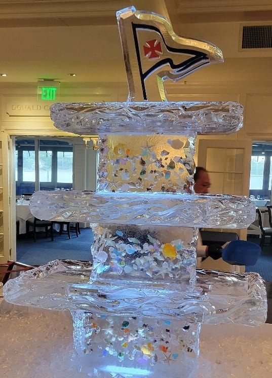 Clear ice sculpture with layered shelves, a small flag on top, and colorful confetti inside a hall
