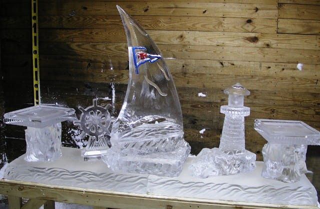 Ice sculptures on a table: a sailboat, lighthouse, wheel, and other frozen objects against a wooden wall.