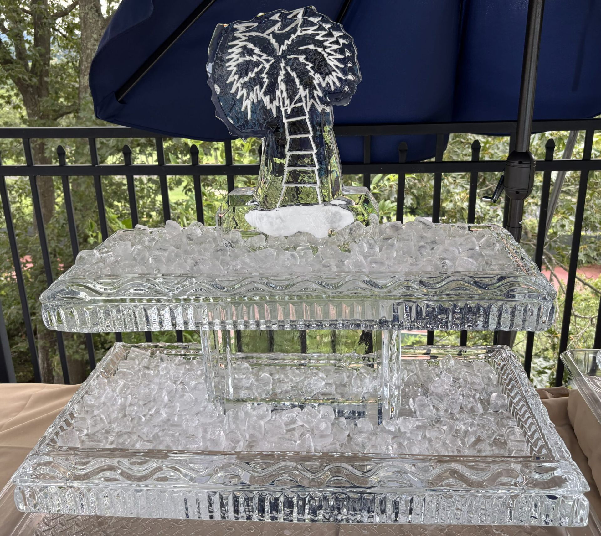 Ice sculpture of a tiered fountain on a patio with a navy umbrella and railing in the background