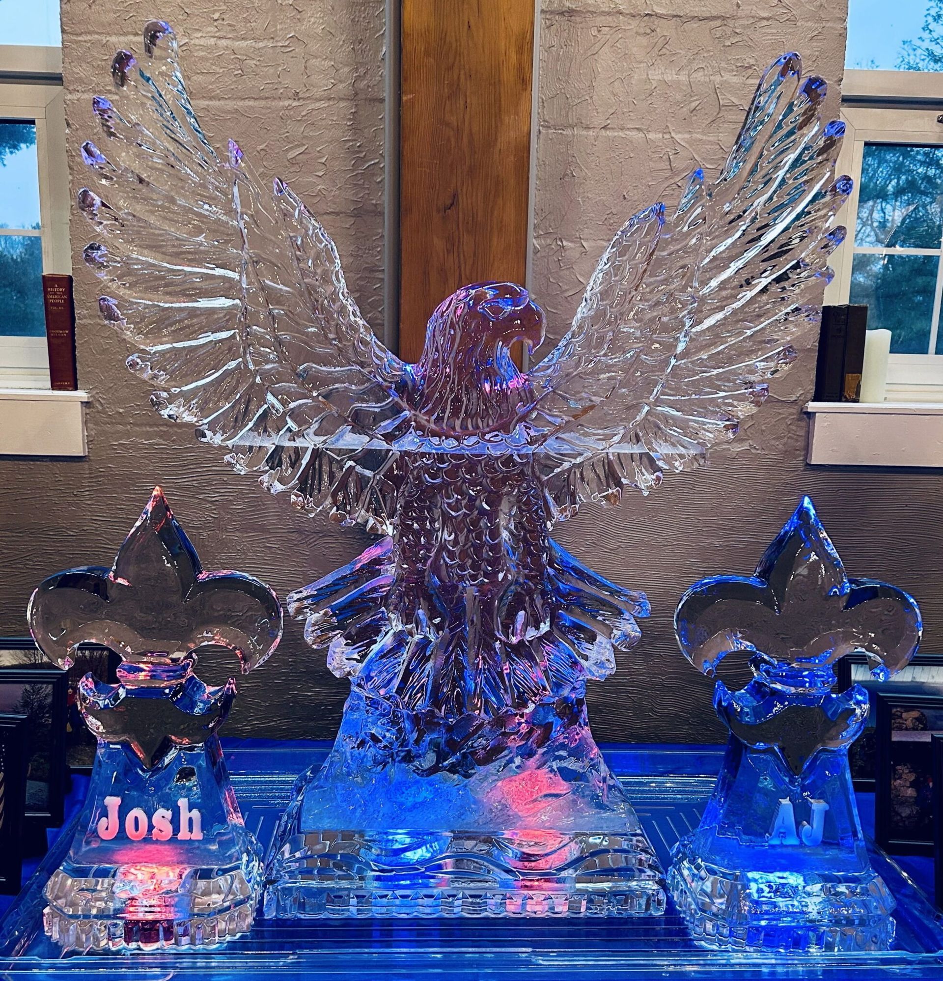 Glowing ice sculpture with outstretched wings on a lit pedestal, flanked by two smaller ice sculptures.