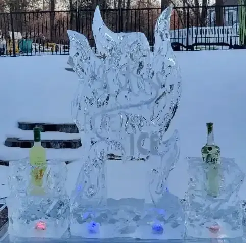 Ice sculpture with glowing lights, flanked by two bottles on a snowy outdoor table.