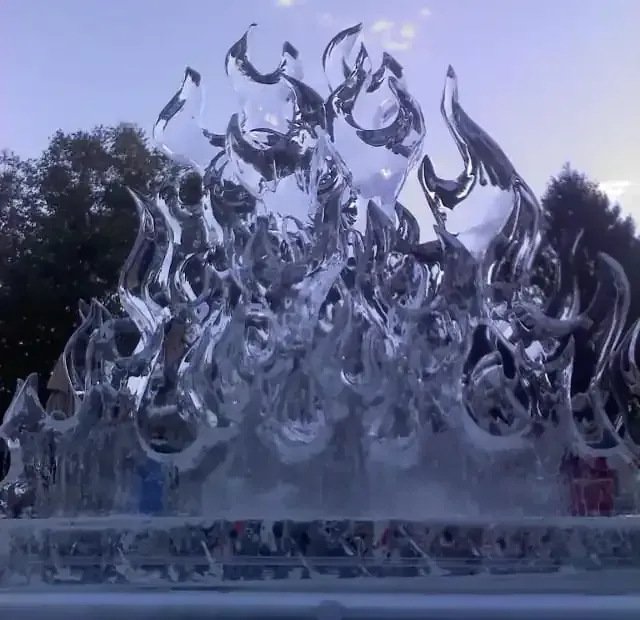 Clear glass sculpture with flame-like shapes in an outdoor setting, against trees and a cloudy sky