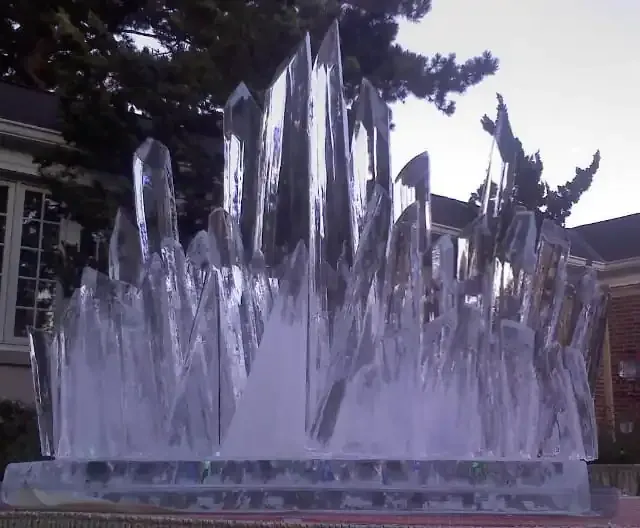 Clear glass sculpture with tall spiky vertical forms outdoors in front of trees and a building