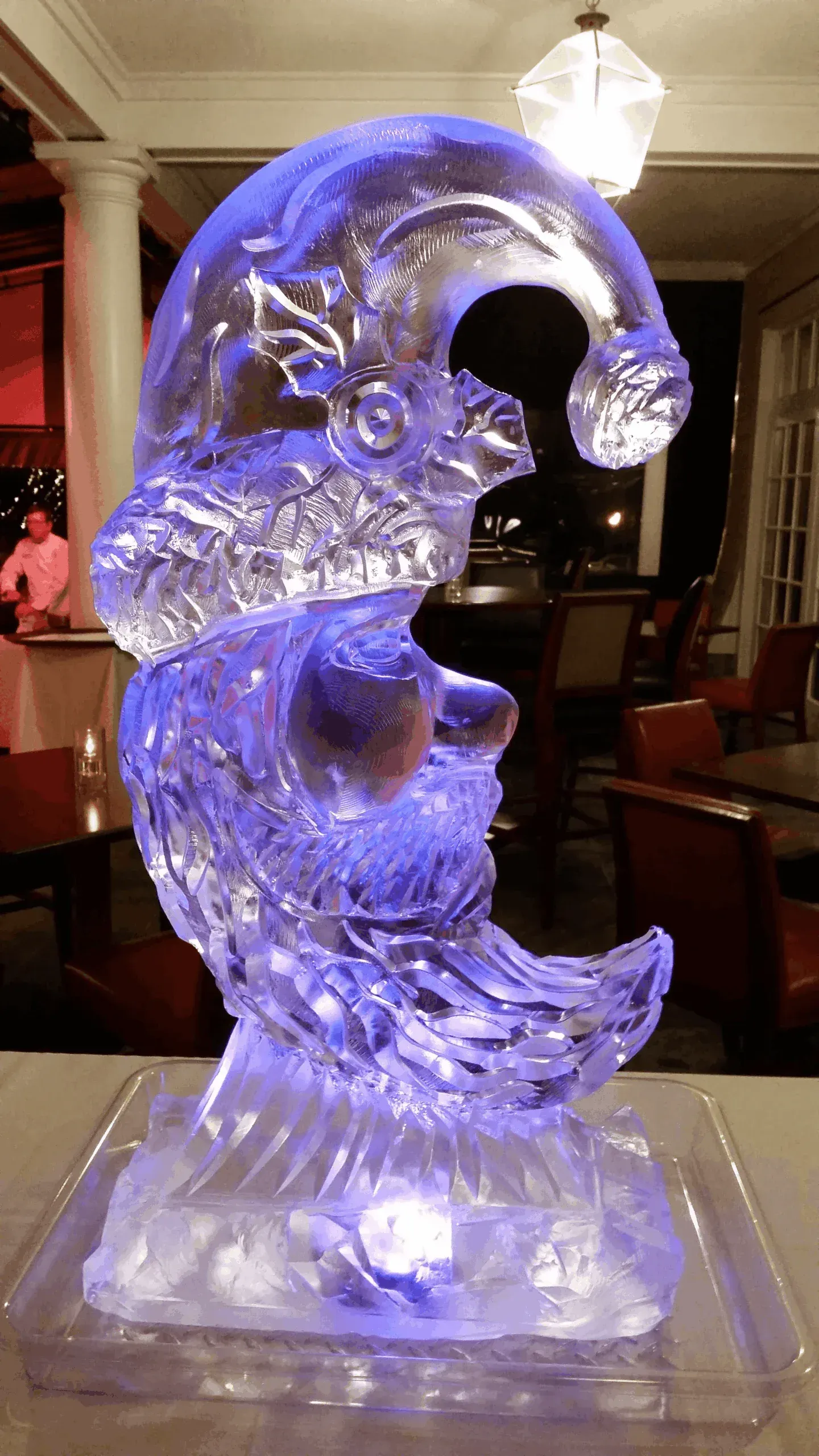 Purple-blue illuminated ice sculpture with a curling wave shape on a pedestal in a dim indoor setting