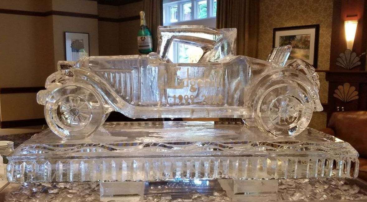 Clear glass sculpture of a vintage car on a reflective pedestal in a warmly lit room