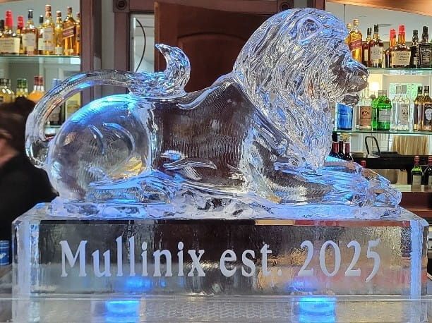 Clear glass lion sculpture on an ice base engraved “Mullinix est. 2025,” displayed at a bar.