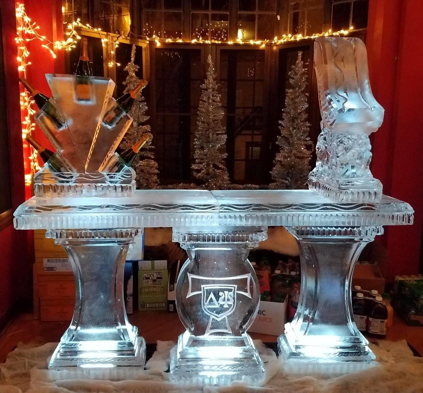 Ice sculpture table display in a warmly lit room, with two sculpted figures and a central etched emblem.