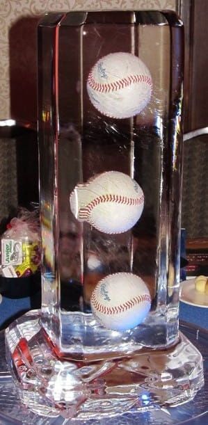 Three baseballs stacked in a clear glass display case on a patterned tabletop.
