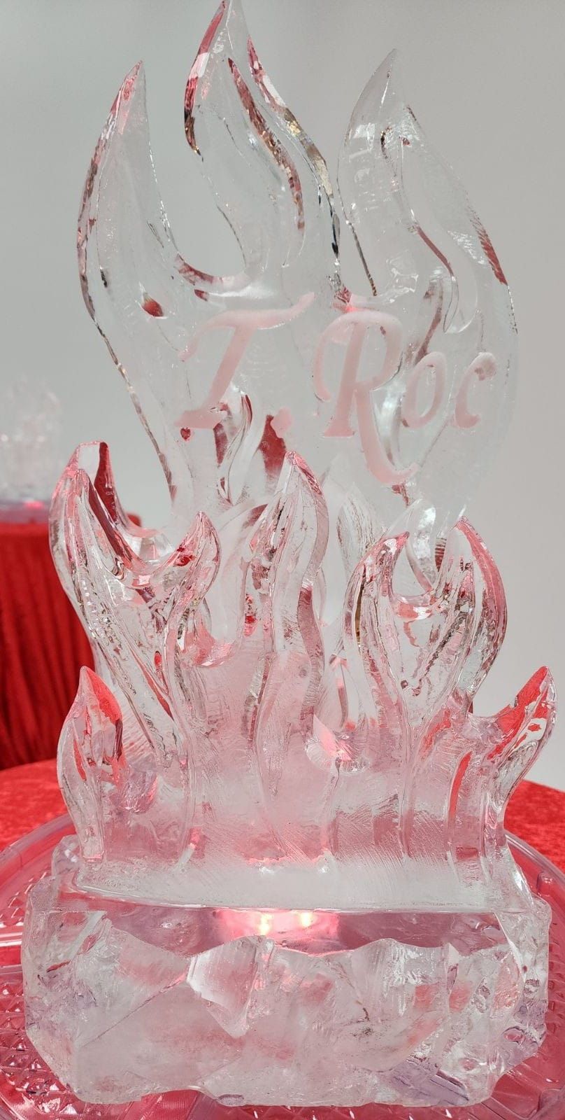 Translucent glass flame sculpture with pink-red accents on a red surface, against a light gray background