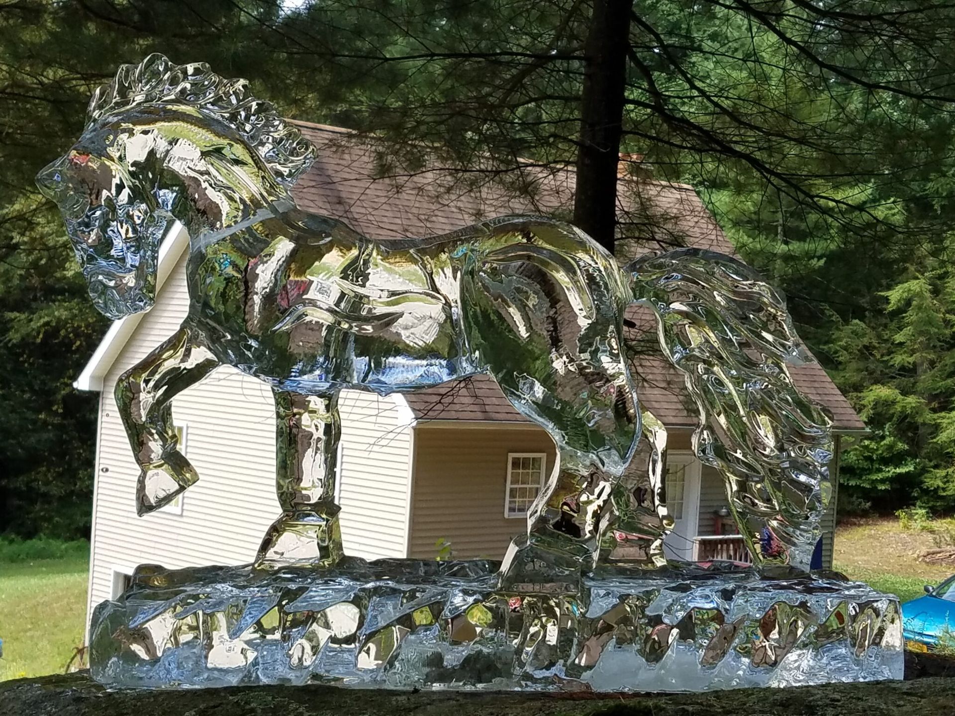 Horse-shaped mosaic sculpture beside a white house, set outdoors among trees.