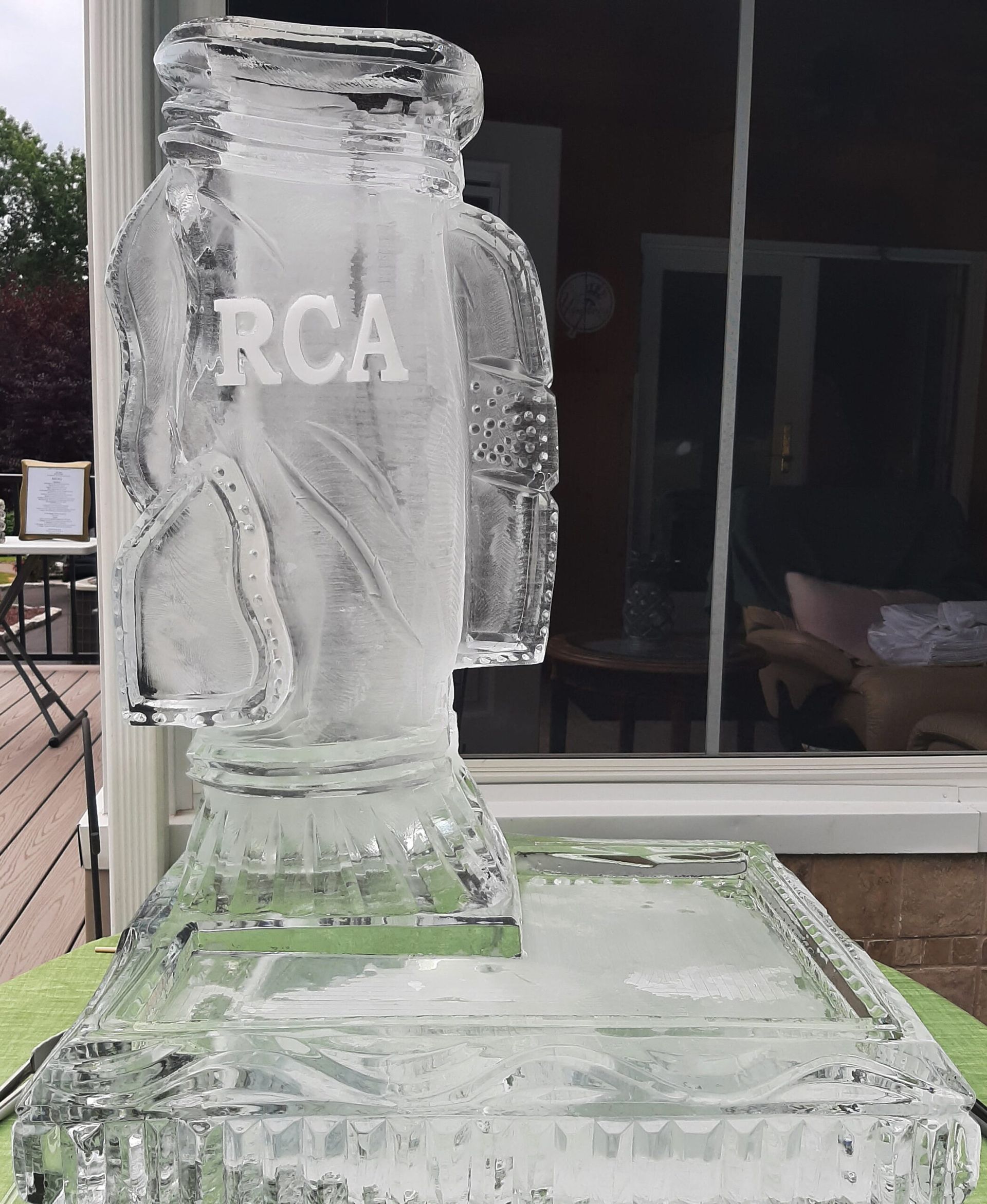 Clear ice sculpture of an RCA bottle on a pedestal outdoors, with a glass door in the background