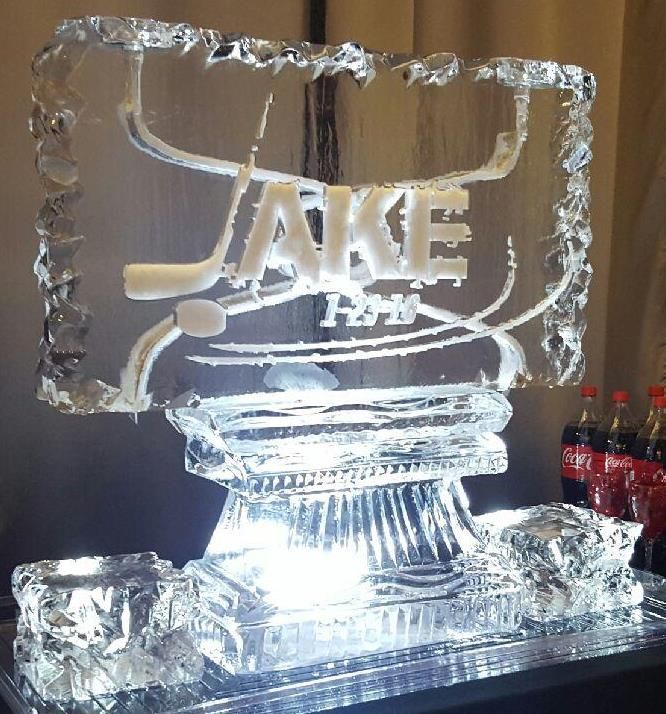 Clear ice sculpture of the word “TAKE” on a lit base, with soda bottles in the background.
