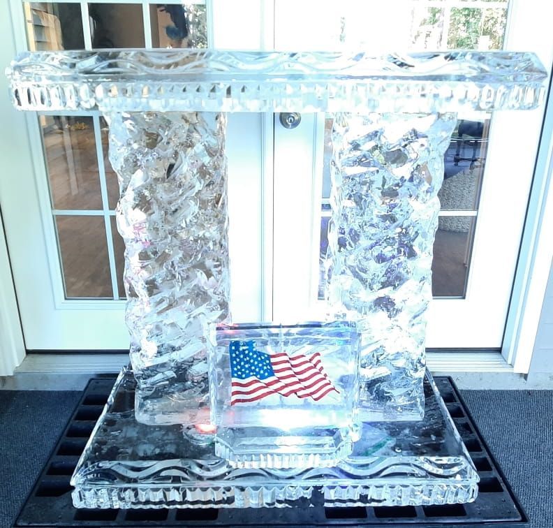 Clear ice sculpture with an American flag centerpiece on a black mat in front of white doors.