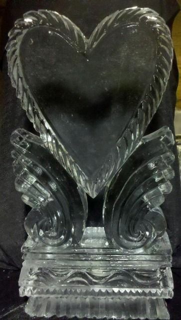 Clear glass heart-shaped ornament with decorative wings and a ridged base, on a dark background