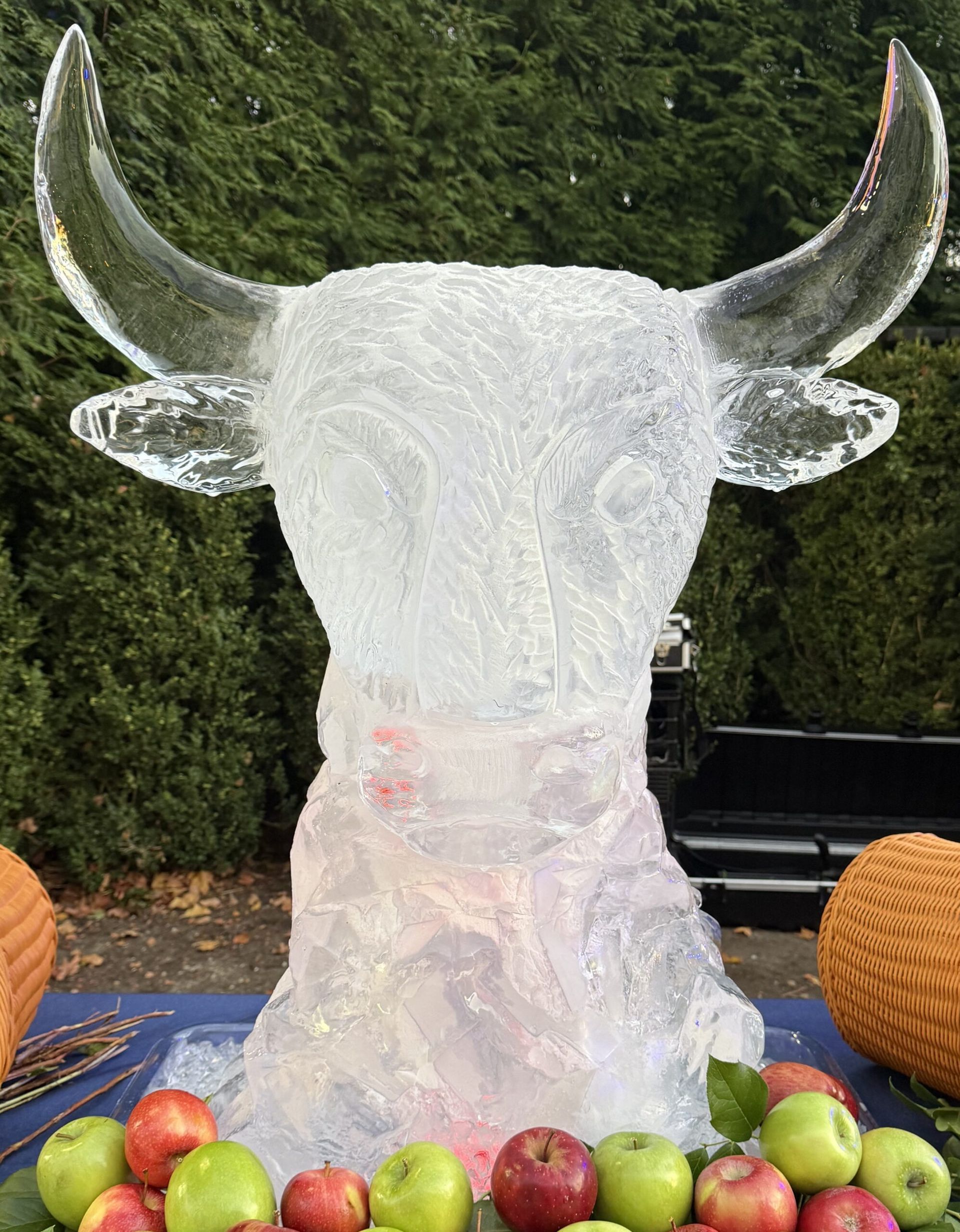 Ice sculpture of a bull’s head with horns, displayed outdoors on a table with fruit in front.