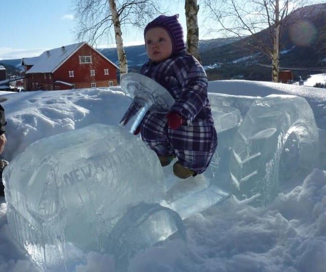 Child in winter clothes sitting on a clear ice sculpture in a snowy yard, with a red house in the background.