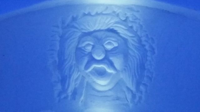 Blue-lit carved face with a haunting expression, centered on a textured wall.