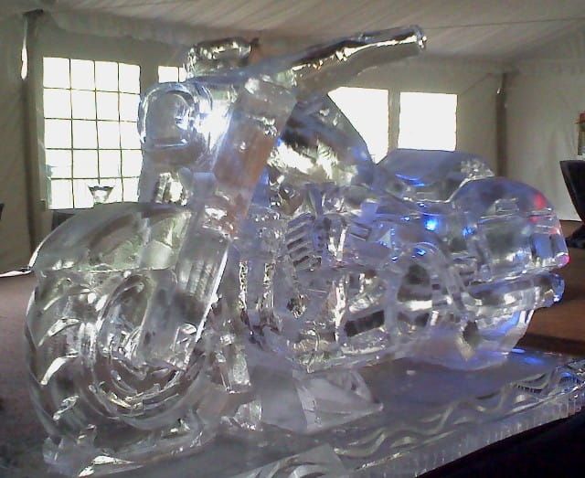 Clear ice sculpture of a motorcycle on a table, lit with bluish reflections in a room with windows.
