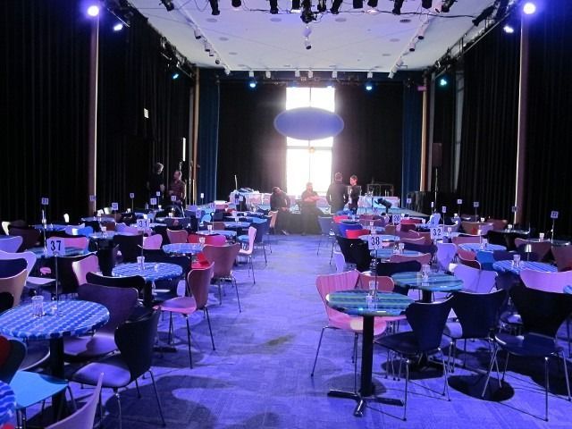 Empty banquet hall with round tables and chairs under purple stage lighting, bright doorway at the far end