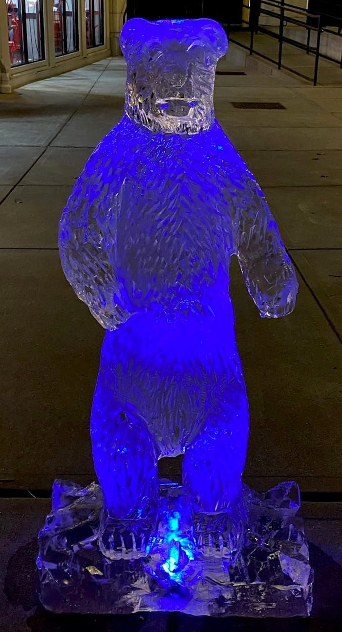 Blue-lit ice bear sculpture standing outdoors at night on a base of ice blocks