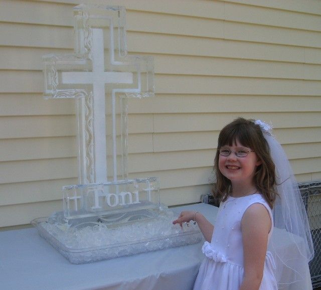 Young girl in a white dress and veil beside an ice cross centerpiece with “Toni” on a table outdoors
