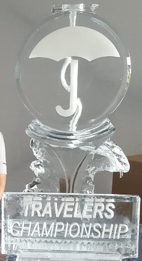 Clear glass Travelers Championship trophy on a reflective base with engraved text.