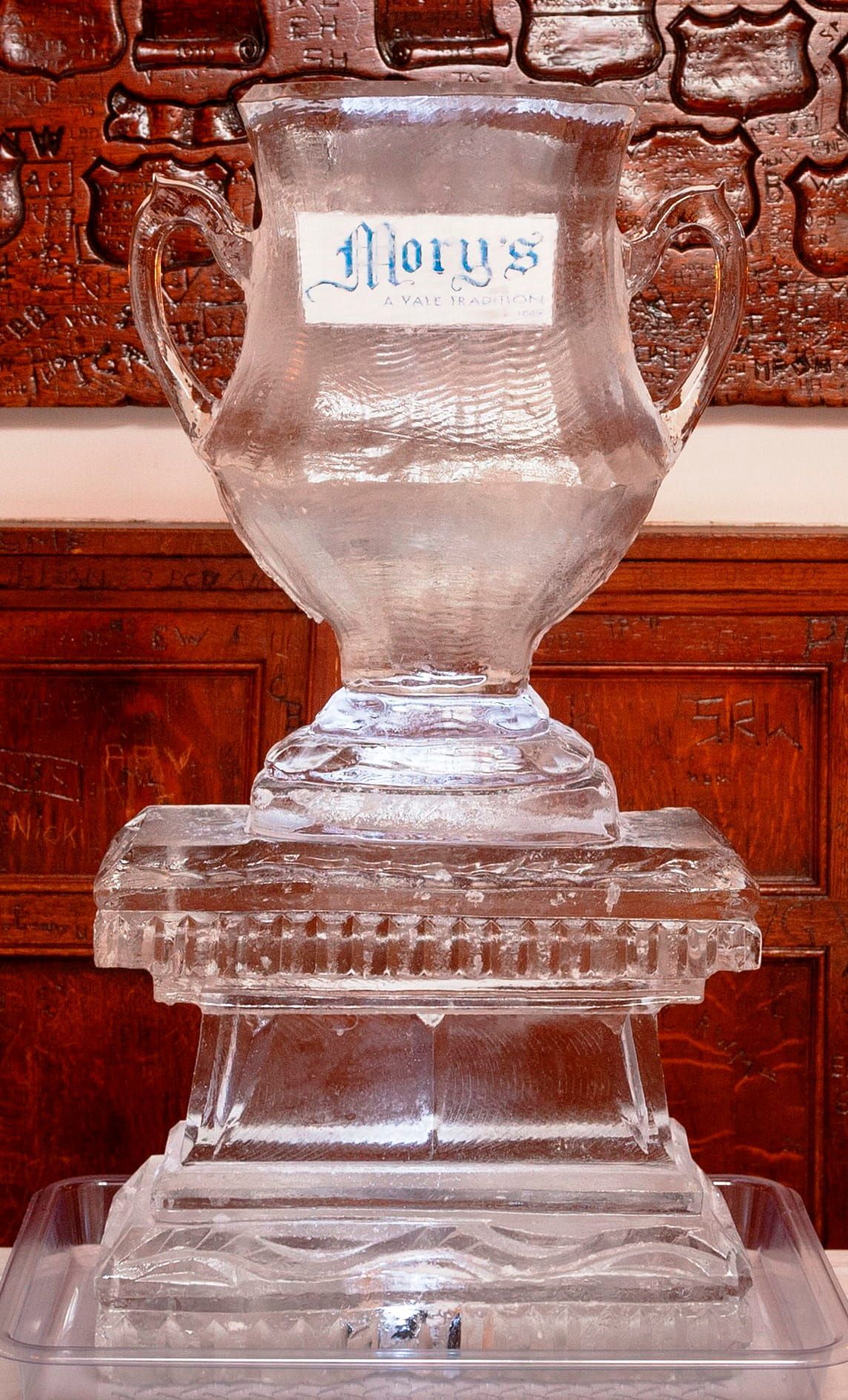 Clear glass urn on a wooden pedestal against a carved brick wall.