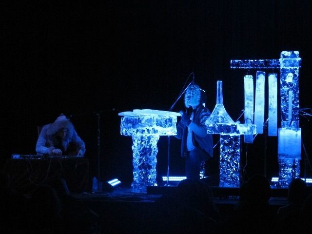 Blue-lit performers on stage beside glowing geometric props and columns in a dark theater