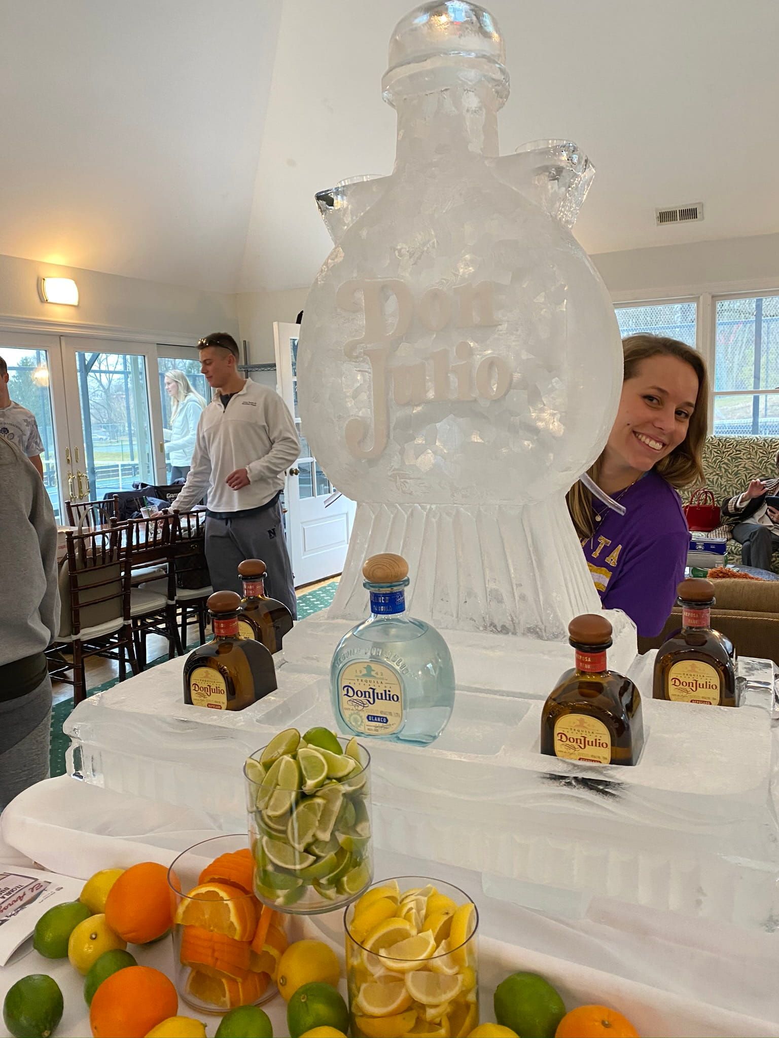 Ice sculpture centerpiece on a dessert table with fruit and bottles in a bright event room