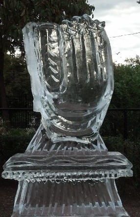 Clear ice sculpture of a crown-like figure on a pedestal outdoors against a dark background