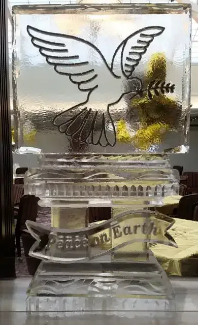 Clear glass memorial block with a white dove and gold accents, resting on a mirrored base with a ribbon reading “on Earth.”
