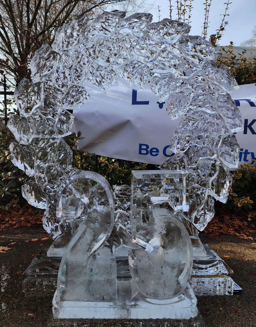 Clear ice sculpture of a tree with two penguins on a base, outdoors near a banner.