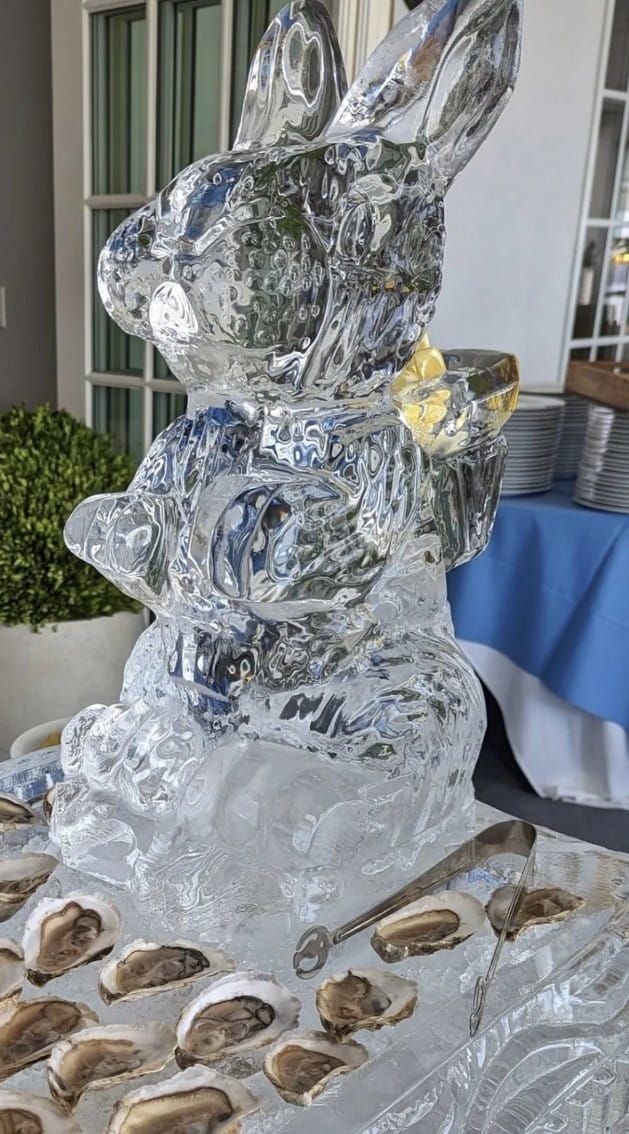 Clear ice sculpture of a rabbit on a table, with seashell decorations and a blue-covered table in the background