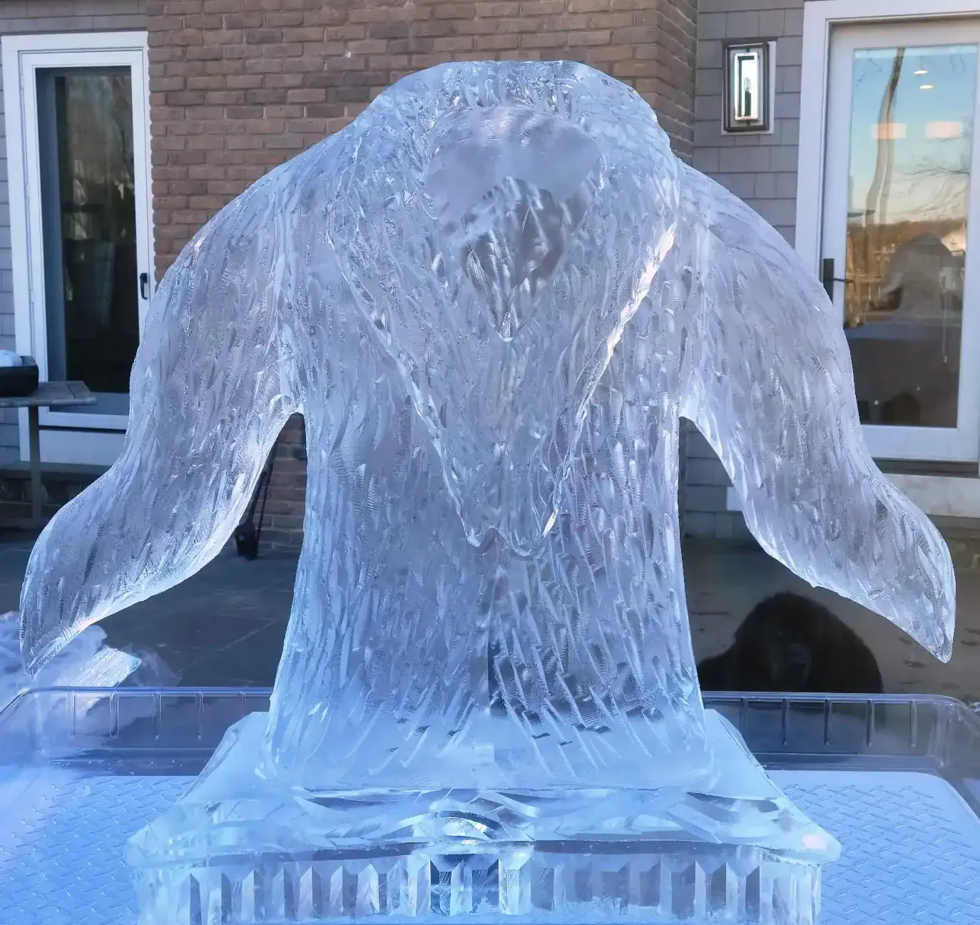 A clear ice sculpture shaped like a penguin standing on a pedestal outdoors in front of a brick building.