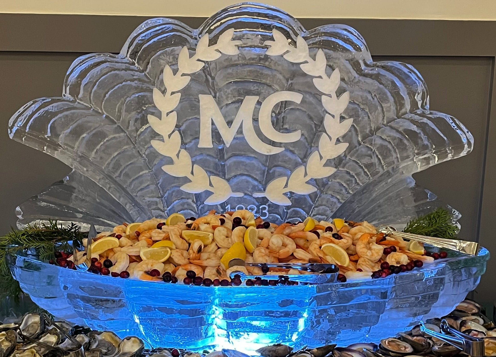 Blue-lit fruit display in a carved ice bowl with MC logo backdrop and laurel wreath design