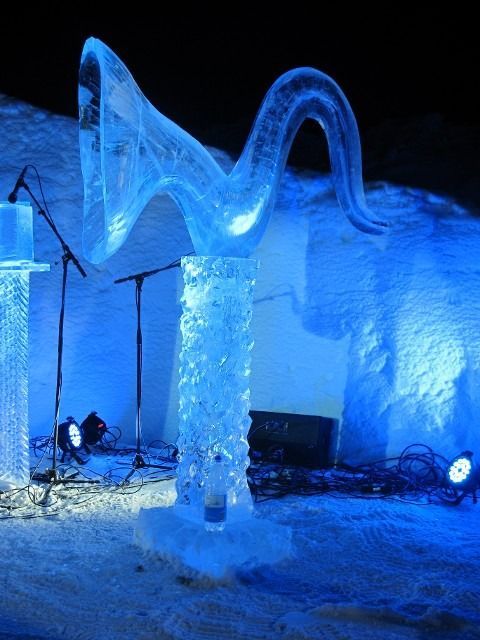 Blue-lit ice sculpture resembling a wave, displayed outdoors at night on snow-covered ground