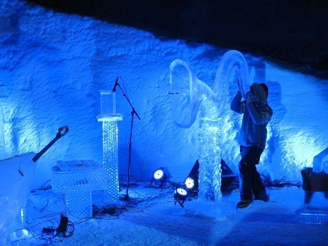 Blue-lit ice sculpture stage with a person playing a trumpet-like instrument beside microphones and speakers