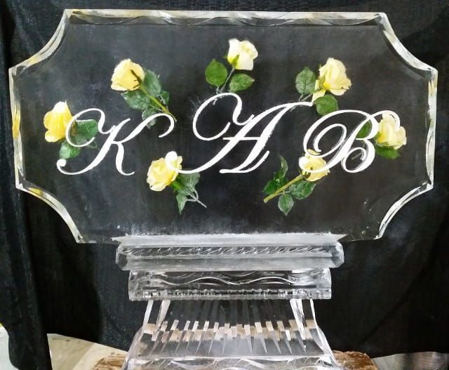 Clear engraved glass plaque with white flowers and script initials “K A B” on a stand