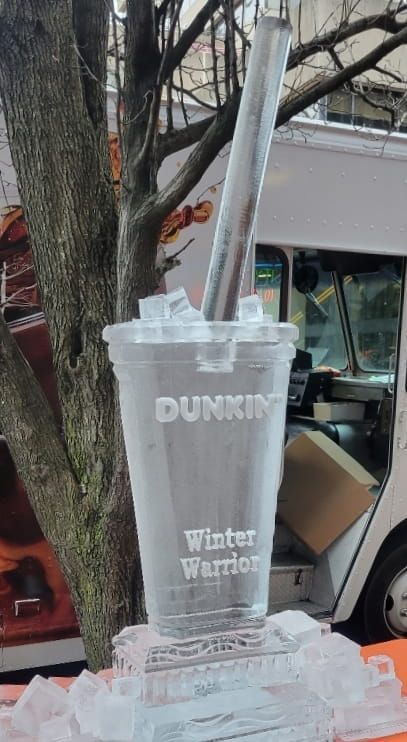 Clear ice sculpture of a Dunkin’ iced drink with straw, outdoors by trees and a storefront.