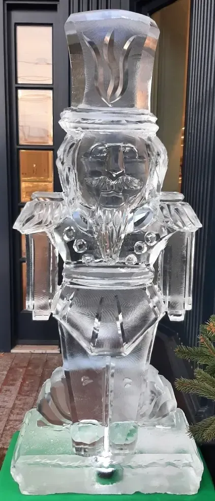 Ice sculpture of a person in a hat on a pedestal, displayed outdoors in front of a building.